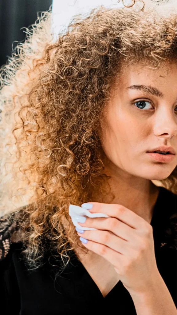 A close-up portrait of a woman with a large volume of tightly textured, curly blonde and light brown hair. She is looking slightly off-camera while using a soft white cotton pad held in her hand, which features light blue painted fingernails. She is wearing a black top, and the lighting highlights the definition of her voluminous curls, serving as a visual example of the tight ringlet results achieved with a heatless scrunchie curling method.