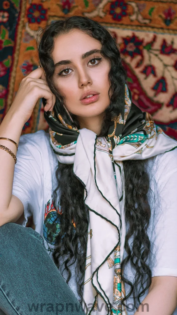 A front portrait of a woman with long, dark, wavy hair sitting in front of a colorful patterned tapestry. She is wearing a cream-colored silk or satin headscarf with dark green and gold ornate borders tied loosely under her chin, letting the long ends drape down the front of her white graphic t-shirt. Her long waves cascade out from underneath the scarf down her shoulders. She leans her head slightly on her right hand, which features gold chain bracelets. The text "wrapnwave.com" is watermarked in grey at the bottom, showcasing how to wear a protective silk scarf over heatless waves.