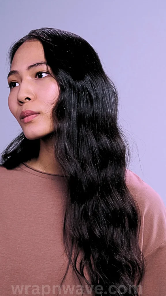 A close-up studio portrait from a three-quarter angle showing a young woman with long, shiny, jet-black hair styled into loose, flowing beach waves. The soft, uniform wave pattern cascades over her left shoulder down past her chest. She is looking off to the side with a serene expression, wearing a plain brown t-shirt against a light lavender-grey background. The text "wrapnwave.com" is watermarked in dark lettering at the bottom, perfectly showcasing the healthy, frizz-free results of a heatless wave routine.