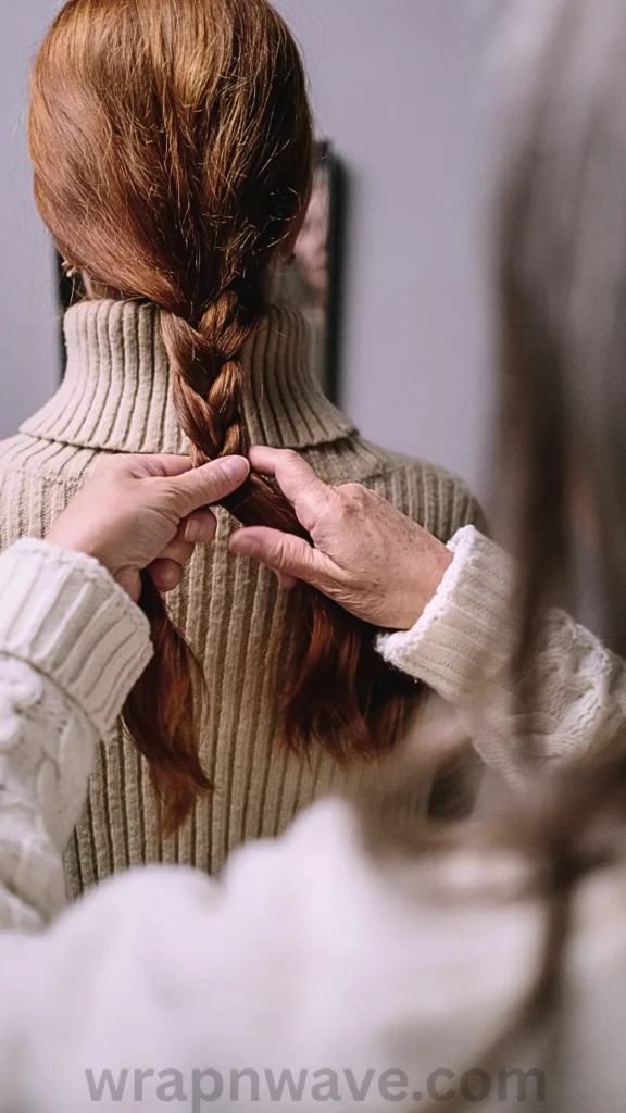 A close-up view from behind of a woman with long, vibrant auburn-red hair. She is being helped by another person (visible as hands and a blurred sleeve in the foreground) who is loosely braiding her hair. The woman is wearing a beige ribbed turtleneck sweater. The hands are actively crossing sections of the braid at the neck level, demonstrating the creation of an overnight braid. A watermark for 'wrapnwave.com' is in the bottom right corner.