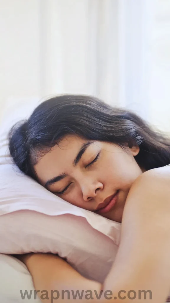 A close-up shot of a young East Asian woman sleeping peacefully on her side in bed. Her eyes are closed with a serene, relaxed expression, and her head is resting on a soft, light pink pillowcase. Her long, dark hair is spread out above her on the pillow as she sleeps. The scene is brightly lit with soft, natural morning light. The text "wrapnwave.com" is watermarked in grey at the bottom, illustrating the effortless overnight process of waking up to heatless waves.