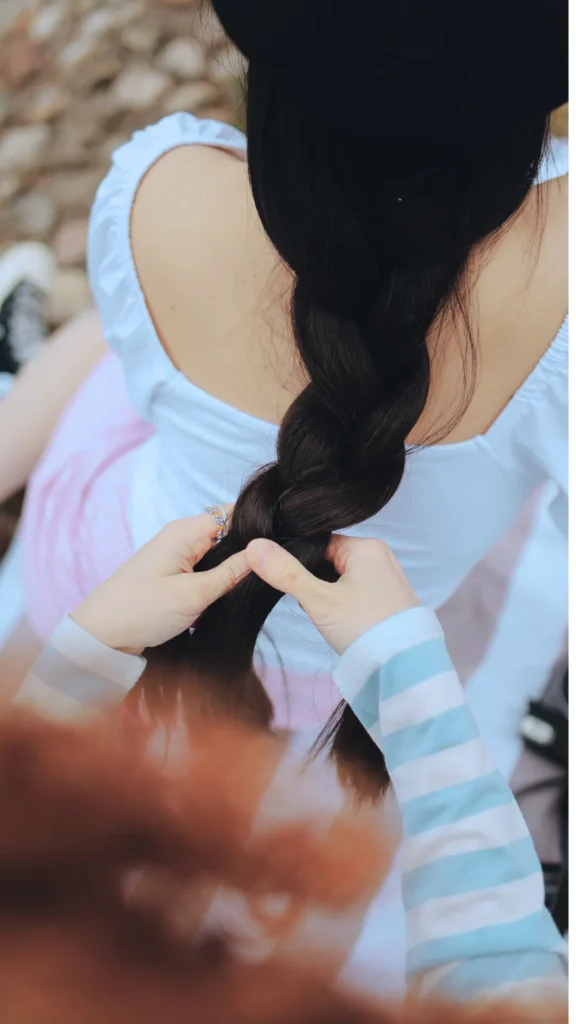 An over-the-shoulder, close-up shot focusing on a person's hands braiding long, thick, dark brown hair. The person braiding wears a light blue and white striped long-sleeve shirt, carefully crossing the hair strands into a classic three-strand braid. The woman receiving the braid is sitting down, wearing a white top and a black hat, seen from behind. The image directly illustrates the step-by-step process of preparing a traditional braid for an overnight heatless wave styling routine.