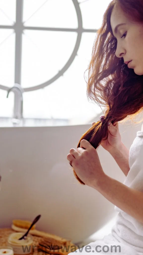 A side-profile view of a woman with long, thick auburn hair sitting in a brightly lit bathroom, looking down with a focused expression. She is wearing a white bath towel or robe and is holding a large section of her wavy hair in both hands, demonstrating how to finger-stretch or separate waves post-styling. In the foreground, out-of-focus bath products, a body brush, and a small container sit on the counter next to a white bathtub under a large circular window. The text "wrapnwave.com" is watermarked in grey at the bottom, providing a visual guide for fixing common styling issues.