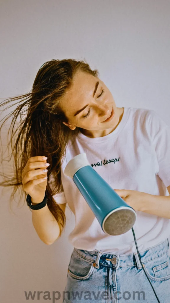 A vertical, medium shot of a young woman rough-drying her hair, as referenced in the file 5 (7).webp.The woman is tilted slightly to her right with her eyes gently closed and a soft smile, holding a vibrant blue and white hair dryer aimed upward at her locks. She uses her left hand to ruffle and lift her long, damp brown hair to build natural texture and volume as the air blows through it. She is wearing a casual white t-shirt with a small green embroidered text design across the chest, light-wash denim jeans, and a black fitness tracker on her left wrist. The shot is set against a plain, off-white background, and a semi-transparent "wrapnwave.com" watermark is visible along the bottom center edge.