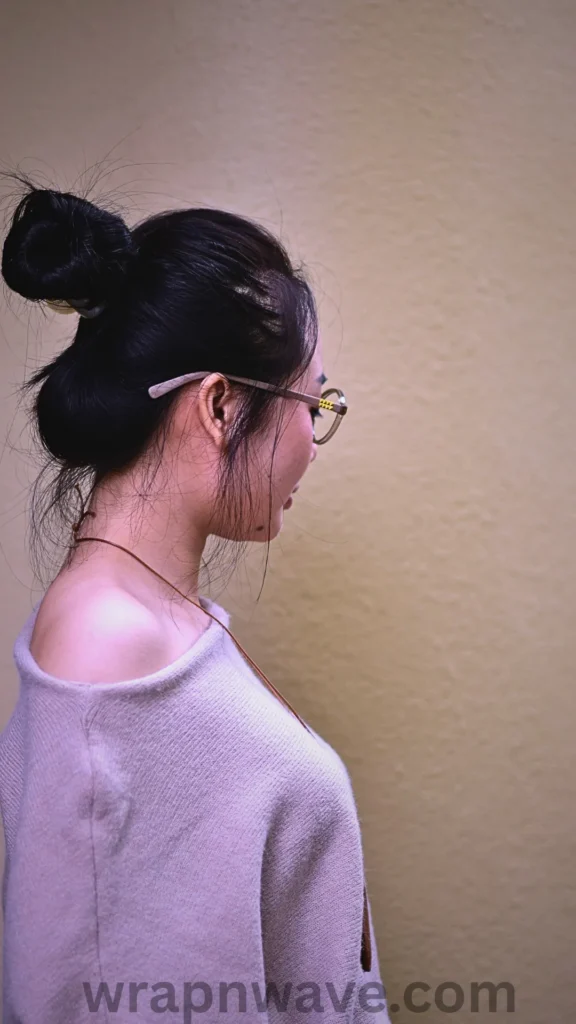 A side profile view of a woman with dark hair styled up into a twisted bun technique on the back of her head, with a few loose strands framing her face and neck. She is wearing round-framed glasses and a light lavender off-the-shoulder knit sweater, posed against a textured, neutral beige wall. The text "wrapnwave.com" is watermarked in dark grey at the bottom, illustrating a comfortable bun setup used to achieve soft, sweeping heatless waves overnight.