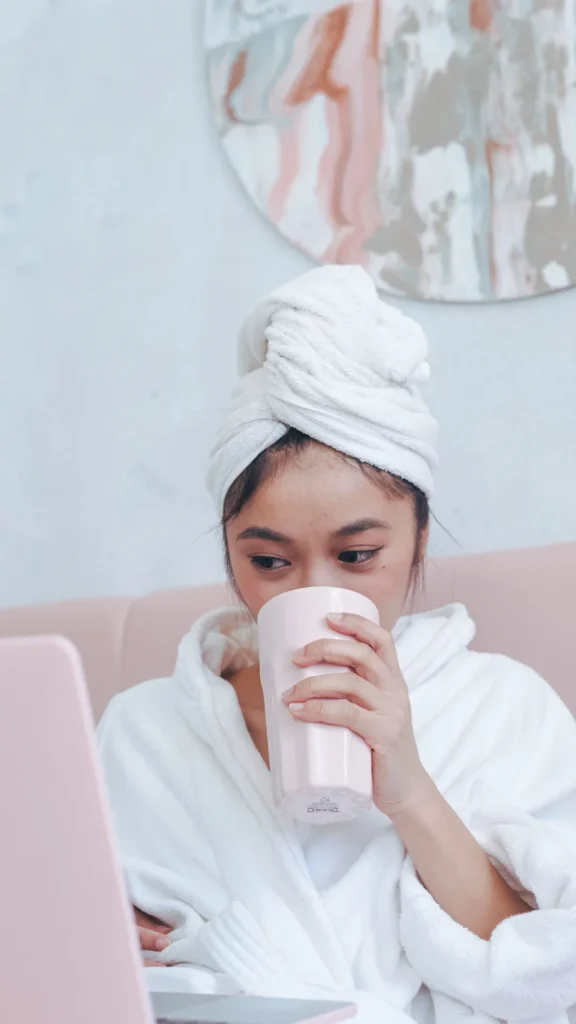 A front view of a young woman sitting comfortably indoors, wearing a plush white bathrobe and a white towel wrapped securely around her wet hair. She is sipping from a tall pink mug held in both hands while looking down at a pink laptop in front of her. The relaxed indoor setting with soft lighting and abstract pink wall art illustrates the prep stage of towel-drying hair for a heatless wave routine.