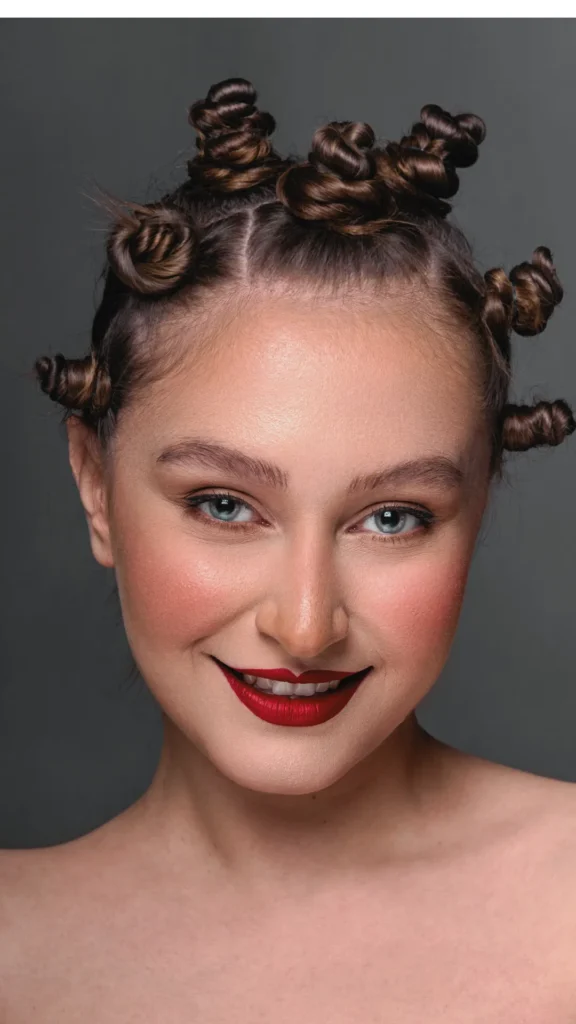 A close-up, front-facing studio portrait of a smiling young woman with light skin and blue eyes looking directly at the camera. Her brown hair is sectioned and styled all over her head into numerous small, tightly coiled, twisted Bantu knots or mini buns. She wears bright red lipstick, rosy pink blush, and has bare shoulders against a solid dark grey studio background. The image provides a clear visual demonstration of creating multiple small buns across the head to achieve tight, defined overnight heatless waves and volume.