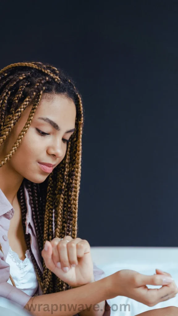 A side-profile view of a young woman with long, neatly styled box braids featuring a mix of dark brown and warm honey-blonde colors. She is looking downward at her hands, appearing relaxed while sitting indoors against a solid dark grey background. She is wearing a white lace-trimmed camisole under a light pink button-down shirt. The text "wrapnwave.com" is watermarked in grey at the bottom, illustrating the protective braided hairstyle used to maintain defined heatless waves.