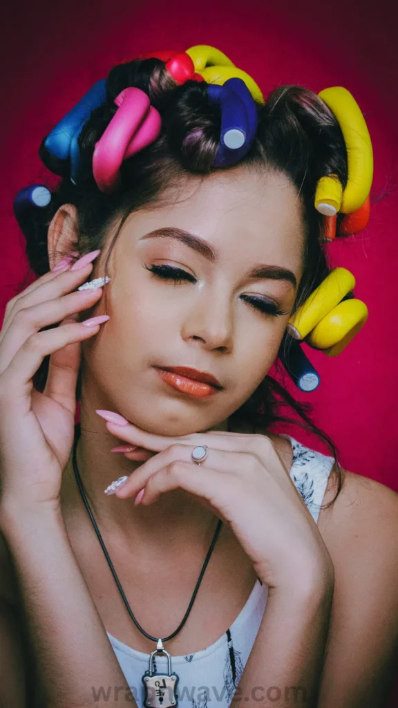 A vertical, close-up photograph demonstrating the placement of multi-colored flexi rods in hair for a heatless curling routine, as referenced in the file Add a heading (26).webp.Shot from a front-facing perspective, the image features a woman posing with multiple bright, flexible foam rollers wrapped tightly throughout her dark hair. The flexi rods are visible around the crown and sides of her head in vivid shades of yellow, pink, blue, and red, twisted into secure shapes to hold the wrapped hair sections.She has her eyes gently closed and hands positioned near her face, showcasing a styled manicure with long, light-pink pointed nails adorned with white floral accents. She is wearing a white patterned top, a thin black necklace with a small padlock pendant, and a silver ring. The portrait is set against a solid, vibrant magenta-pink studio background. Along the bottom center edge, a clean, dark-grey watermark reads "wrapnwave.com".