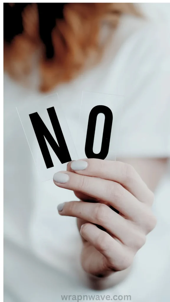 A close-up shot of a person's hand holding up two small, clear plastic cards that display the word "NO" in bold black block letters. The person has a light grey manicure and is wearing a white top, with their torso and light brown hair softly blurred in the background. The text "wrapnwave.com" is watermarked in grey at the bottom. The image serves as a visual cue for a hair styling "don'ts" or troubleshooting section, illustrating common mistakes to avoid when wrapping heatless waves.