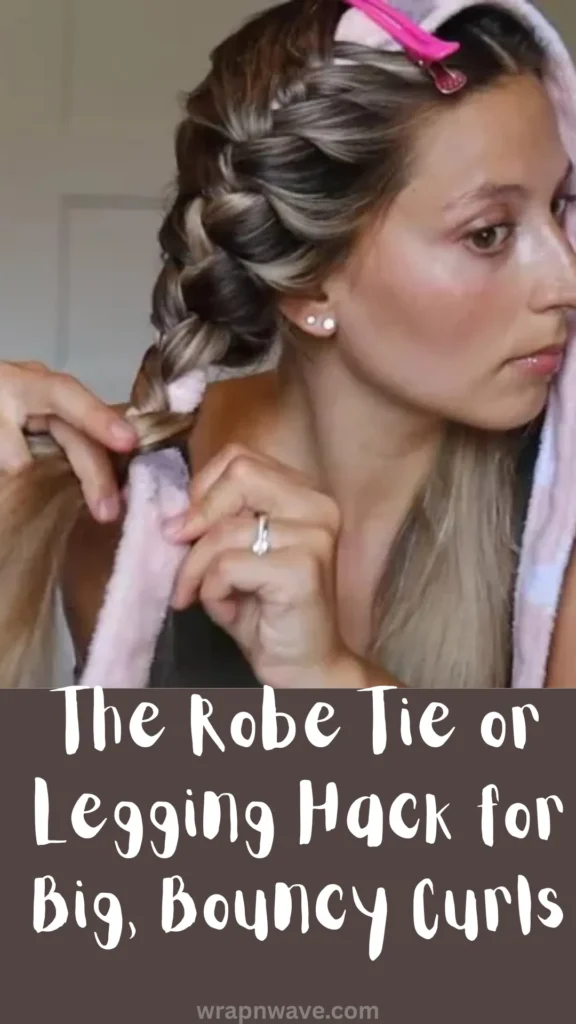 A vertical graphic design showcasing a tutorial step for heatless hair styling, as referenced in the file The Robe Tie or Legging Hack for Big, Bouncy Curls.webp.The top half of the image features a close-up photograph of a young woman with blonde-highlighted hair demonstrating the robe tie curling method. A plush pink and white robe tie is placed over the top of her head, secured at her crown with a bright pink sectioning hair clip. She uses both hands to tightly wrap and braid a thick section of her hair down the side of the robe tie.The bottom half of the image features a solid, dark brown rectangular text block with casual, handwritten-style white text that reads, "The Robe Tie or Legging Hack for Big, Bouncy Curls". The website URL "wrapnwave.com" is printed in small, muted grey text at the very bottom center edge.