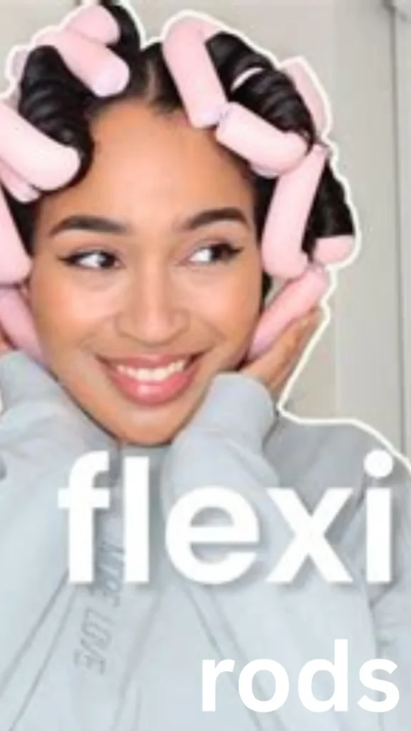 A vertical, close-up photograph demonstrating the use of heatless flexi rods for hair styling, as referenced in the file rods (1).webp.The image features a smiling woman with a center part looking to her right while gently resting her hands against her cheeks. Her dark hair is sectioned and tightly wound around several thick, light pink foam flexi rods that encircle her head. She is wearing a light grey crewneck sweatshirt. The woman has a thin white outline around her silhouette against a blurred background. Large, bold white text is overlayed across her chest area, reading "flexi rods".
