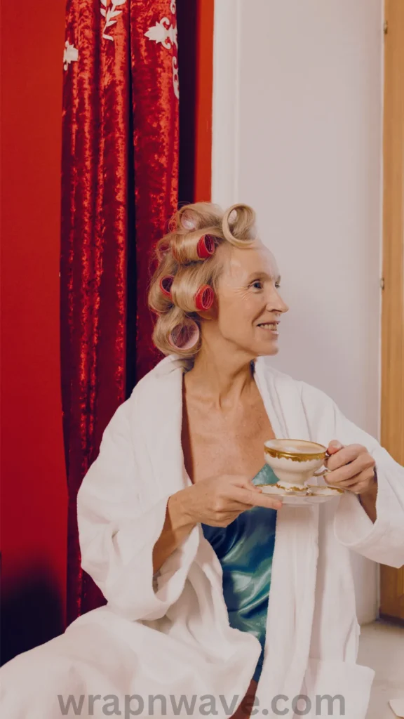 A medium, three-quarter portrait of an older woman with blonde hair looking to the side with a warm smile. Her hair is wrapped in large pink and red vintage hair rollers to set curls. She is relaxed, wearing a white plush bathrobe over a teal satin slip dress, and holding a classic porcelain teacup and saucer. The background features a dramatic red velvet curtain and a white door frame. The text "wrapnwave.com" is watermarked in grey at the bottom, perfectly illustrating a comfortable morning routine while waiting for heatless waves or rollers to set.