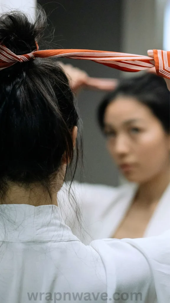 A close-up shot from behind a woman with dark hair as she stands in front of a mirror, preparing an overnight heatless styling routine. She is wrapping a burnt-orange and white striped fabric tie or scarf over the crown of her head. Her blurred reflection in the mirror shows her looking forward while adjusting the fabric with her hands. She is wearing a white robe or shirt. The text "wrapnwave.com" is watermarked in grey at the bottom, providing a clear demonstration of the robe tie curl setup.