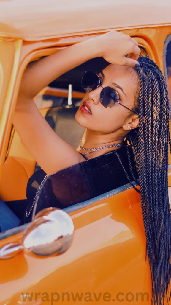 A close-up profile portrait of a woman leaning out of the open window of a bright orange vintage car. She has long, dark micro braids with lighter brown highlights that cascade over her shoulder down past the car door. She is wearing round black sunglasses, red lipstick, a black top, and a silver chain necklace, looking down toward the camera with her right arm raised to touch her head. The text "wrapnwave.com" is watermarked in grey at the bottom, perfectly illustrating how multiple small, tight braids are worn before being unraveled for defined heatless waves.