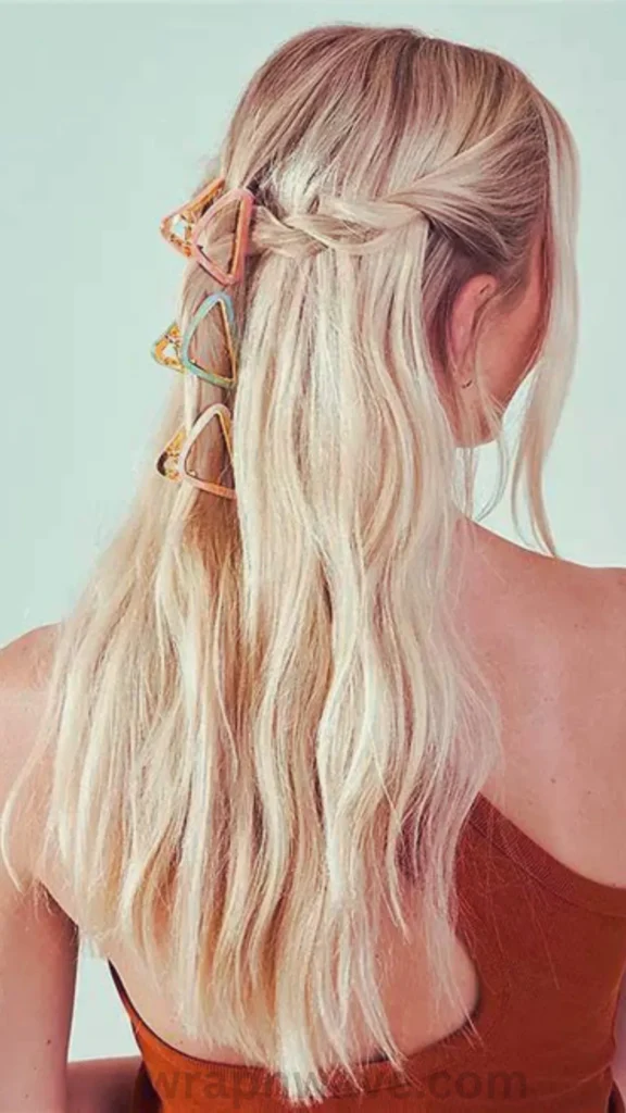 A rear close-up view of a woman with long, textured platinum blonde hair with light pink undertones, styled in loose, air-dried summer waves. The top sections of her hair are loosely pulled back into a delicate half-up twist, which is secured down the center with a vertical stack of three small, triangular geometric claw clips in pastel shades of pink, blue, and orange with gold borders. She is wearing a brown tank top against a plain, off-white background. The text "wrapnwave.com" is watermarked in dark grey at the bottom, illustrating a fresh and easy way to accessorize summer waves.