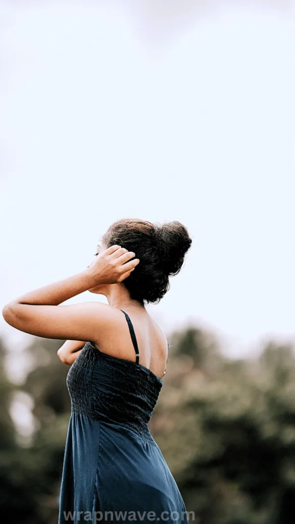 A full-body outdoor portrait of a woman with long, dark, wavy hair pulled up into a high, textured messy bun, demonstrating the final coiled hairstyle. She is standing with her back to the camera, looking to the left with her right hand raised to adjust her bun and her left arm bent. She is wearing a dark, smocked spaghetti strap sundress. The background features a soft-focus forest edge under a bright, high-key white sky, illustrating the results of a comfortable twisted-bun heatless styling technique. The text "wrapnwave.com" is watermarked in grey at the bottom.