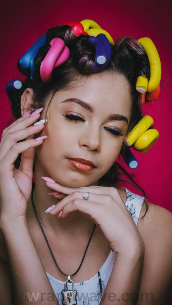 A portrait of a woman with her eyes gently closed, posing against a vibrant magenta background with multi-colored flexi rods set in her hair. Her hair is wrapped around various brightly colored foam rods including yellow, pink, blue, and purple, demonstrating how different rod sizes and colors are used for heatless styling. She has long pink manicured nails, light makeup, and wears a padlock pendant necklace. The text "wrapnwave.com" is watermarked in grey at the bottom.