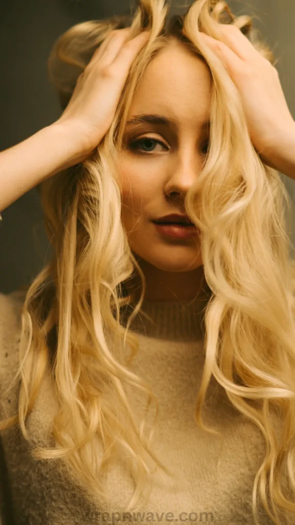 Here are a few ALT text options for the image wrapnwave.com (56).webp, written to match the context of your "lived-in" twist-out wave tutorial:Option 1: Detailed & Context-Focused (Recommended)A close-up portrait of a young woman with long, voluminous, golden blonde hair styled in soft, textured waves. She has her hands gently framing her forehead and temple as some wavy blonde strands casually fall across her face. She has light eyes and is wearing a cozy, beige knit sweater against a moody, dark grey background. The text "wrapnwave.com" is watermarked in grey at the bottom, showcasing the voluminous, "lived-in" results of a heatless twist-out routine.Option 2: Concise & DirectFront view of a blonde woman running her hands through her long, wavy blonde hair. She is wearing a warm beige sweater against a dark background, illustrating the textured and bouncy waves achieved from a heatless twist-out method. Includes a "wrapnwave.com" watermark at the bottom.Option 3: Ultra-Short (Under 125 characters)Woman with long, wavy blonde hair styling it with her hands. Includes a "wrapnwave.com" watermark at the bottom.