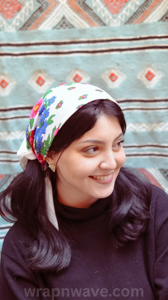 A close-up, three-quarter portrait of a smiling woman with dark, wavy hair looking off to the side. She is wearing a white silk headscarf decorated with vibrant blue, pink, and green floral patterns tied over her head like a bandana. She is dressed in a black turtleneck sweater and small triangle earrings, posed against a textured textile background with geometric diamond patterns in muted teal, cream, and red. The text "wrapnwave.com" is watermarked in grey at the bottom, showcasing a stylish way to protect or set hair for a heatless headband routine.