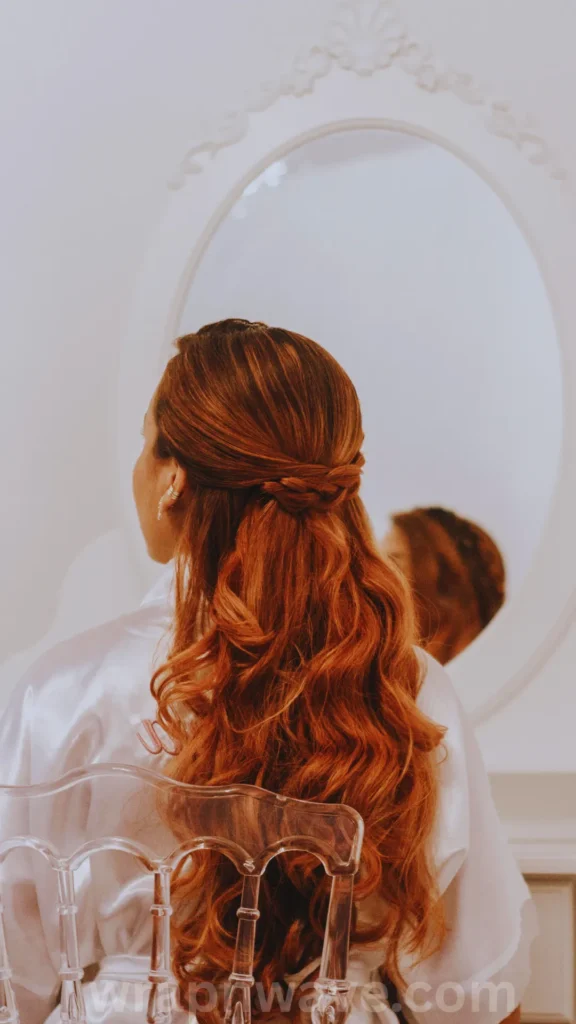 A vertical, close-up photograph captured from behind a woman with long, vibrant auburn hair, demonstrating a damage-free, heatless hairstyle, as referenced in the file wrapnwave.com (75).webp.The woman is seated in a transparent acrylic chair facing an ornate, white oval vanity mirror, showing her back and a slight profile of her face. Her rich reddish-brown hair is styled in a half-up layout, with sections from the sides elegantly twisted and pinned together at the back of her head, while the rest of her thick, long hair cascades down her back in loose, voluminous waves. She is wearing a silky white satin robe. The setting is bright and brightly lit, with a faint, semi-transparent "wrapnwave.com" watermark running along the bottom center edge.