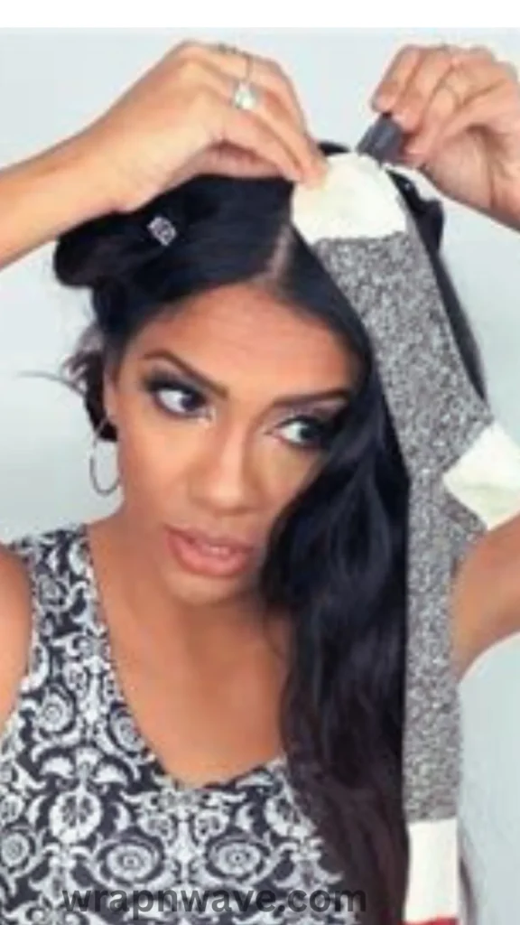 A vertical, close-up photograph demonstrating the heatless "sock curls" hair styling method, as referenced in the file wrapnwave.com (76).webp.The image shows a woman with long, dark brunette hair split down a neat center part. She is looking slightly off-camera while using both hands to anchor a long, patterned grey and cream-colored sock to the top of her head near her roots with a small hair clip. The rest of the sock hangs down the left side of her face, ready for sections of hair to be wrapped around it. She is wearing large silver hoop earrings and a black and white patterned sleeveless top. A dark, semi-transparent "wrapnwave.com" watermark runs along the bottom center edge against a plain white background.