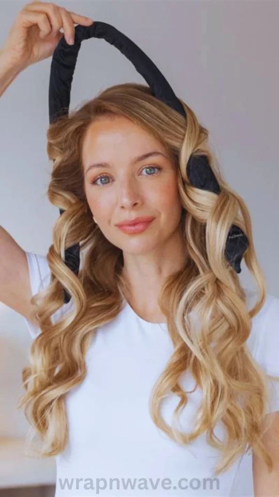 A front portrait of a smiling woman with long blonde hair demonstrating the removal of a heatless hair styling tool. She is using her right hand to lift a long, U-shaped black satin curling ribbon off the top of her head. Her hair falls in voluminous, beautifully defined spiral waves over her shoulders where it has been unrolled from the tool. She is wearing a simple white t-shirt against a plain grey background. The text "wrapnwave.com" is watermarked in grey at the bottom, showcasing the immediate results of a damage-free overnight wave routine.