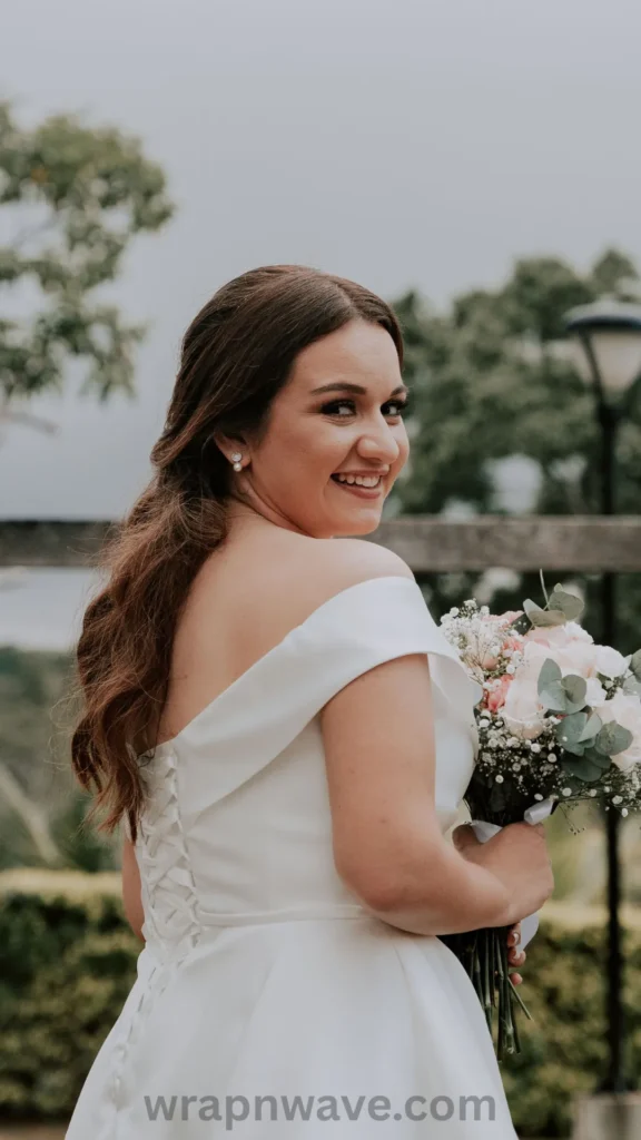 A vertical, close-up bridal photograph capturing a finished wedding hairstyle with soft, heatless waves, as referenced in the file wrapnwave.com (99).webp.Shot from a rear three-quarter angle, the image features a smiling bride looking back over her right shoulder directly toward the camera. Her medium-long, chestnut-brown hair is half-swept back, letting loose, textured waves flow down her upper back. She wears elegant pearl stud earrings and an off-the-shoulder white satin wedding dress featuring a classic lace-up corset back.She is holding a bridal bouquet of white and blush pink roses mixed with green eucalyptus leaves. The background shows a softly blurred outdoor setting with a wooden railing, green trees, and an overcast sky. Along the bottom center edge, a clean, dark-grey watermark reads "wrapnwave.com".