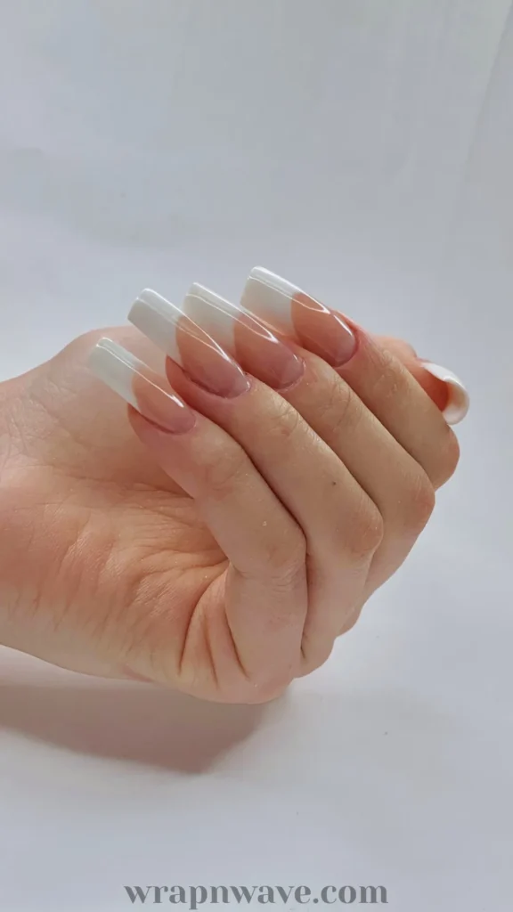 A hand displaying long, square-shaped acrylic or gel nails styled in a classic French tip design, featuring a glossy nude base and clean white tips against a plain light background. An elegant visual example for modern French tip nail trends and manicures.