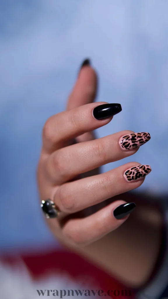 Close-up of a hand with long coffin-shaped nails featuring a high-gloss black polish mixed with pink leopard print accent nails. Set against a soft blue background, this trendy animal print manicure showcases bold gel nail art ideas.