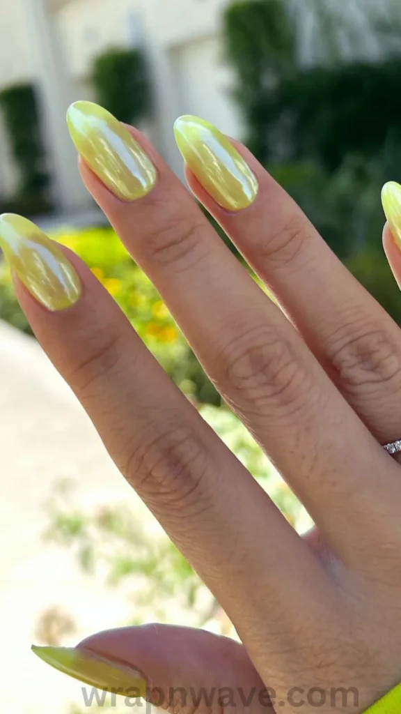 Close-up of a hand with long, almond-shaped gel nails featuring a vibrant chartreuse green base with a glossy glazed donut chrome finish, shown outdoors in bright sunlight. A colorful visual representation of modern glazed donut nail trends and iridescent chrome manicures.