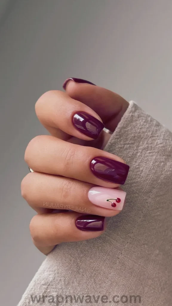 Close-up of a hand wearing a cozy beige knit sweater, showcasing short square gel nails in glossy deep burgundy with a light pink ring finger accent featuring tiny painted red cherries. A stylish visual example for versatile, chic gel nail designs.
