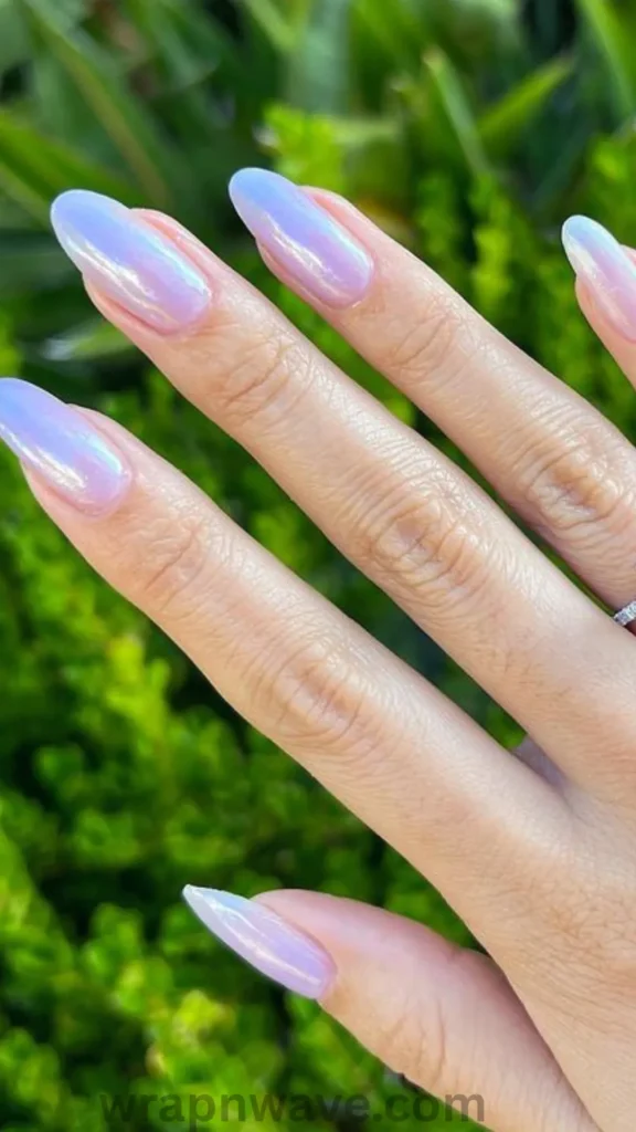 A hand with long, almond-shaped gel nails featuring a sheer lavender-pink base topped with an iridescent chrome glazed donut finish, shown outdoors against a blurred green leaf background. A perfect visual representation of glazed donut nails and chrome powder gel manicures.