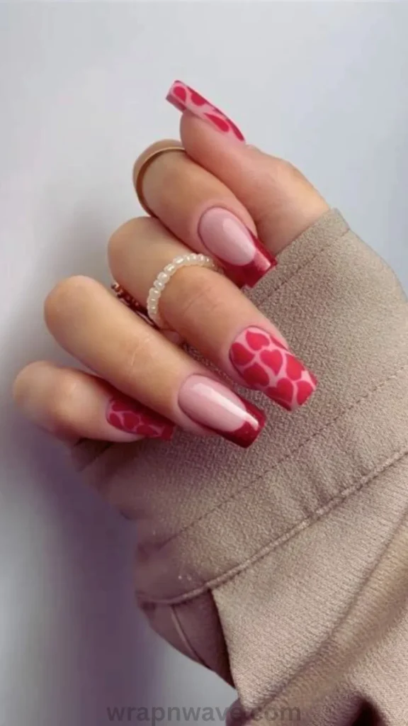 Close-up of a hand with long square nails wearing gold rings, featuring a nude base styled with red French tips and red heart pattern accent nails, tucked into a beige sleeve. A creative visual example for trendy gel nail art designs.