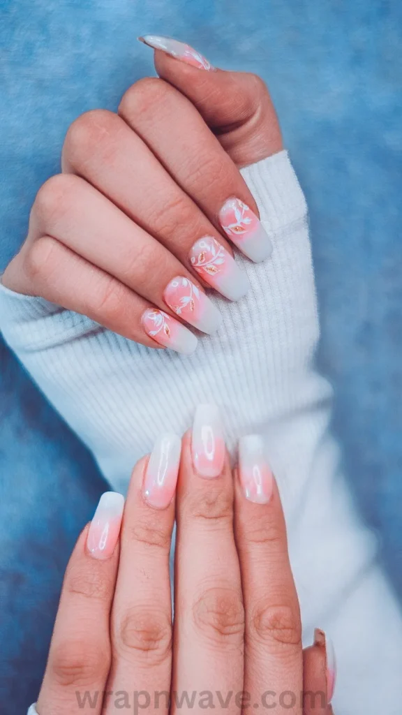 Close-up of two hands featuring long square gel nails with a pink and white gradient ombre base. The top hand showcases delicate white floral vine nail art with gold foil leaf accents, worn over a white ribbed knit sleeve against a blue background. A chic visual example of floral nail art and botanical manicure trends.