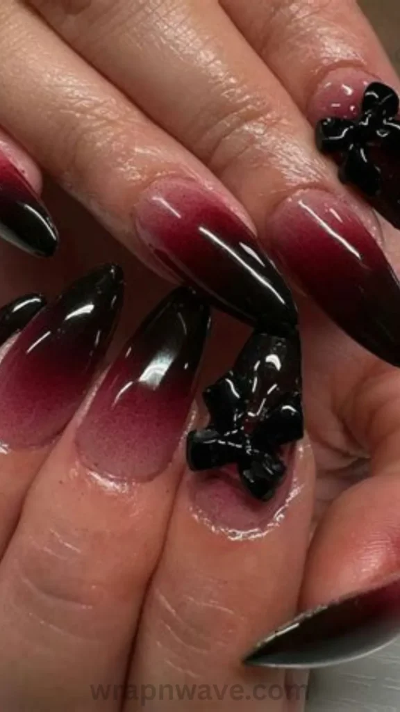 Close-up of long almond gel nails featuring a glossy ombre gradient fading from deep burgundy to black, decorated with bold black 3D bow charms. A striking visual example of dark coquette nail art and edgy fall manicures.