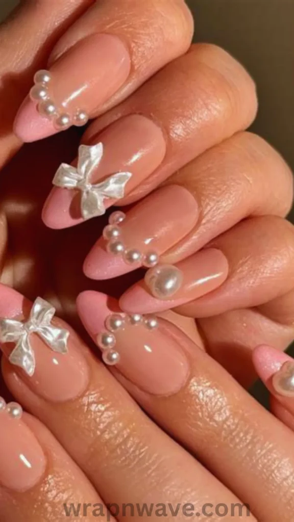 Close-up of almond-shaped gel nails featuring a sheer nude base with soft pink French tips, decorated with 3D white fabric bow charms and delicate pearl studs along the French tip smile lines. A visual example of pearl bow coquette nail art and wedding manicure trends.