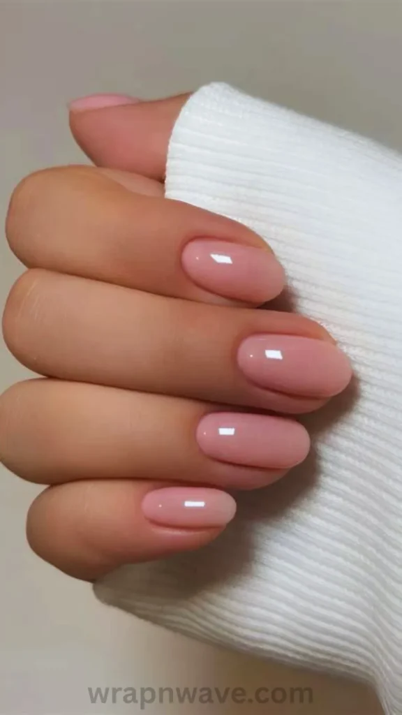 Close-up of short oval nails painted in a sheer, glossy strawberry milk pink gel polish, tucked gently into a white ribbed sweater sleeve. A clean visual example of strawberry milk nails and minimal coquette manicures.