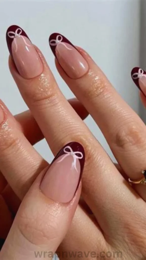 Close-up of almond-shaped gel nails featuring a sheer nude base with deep burgundy French tips, decorated with delicate white painted ribbon bows and tiny pearl dots centered on each tip. A striking visual example of French tip bow nail art.