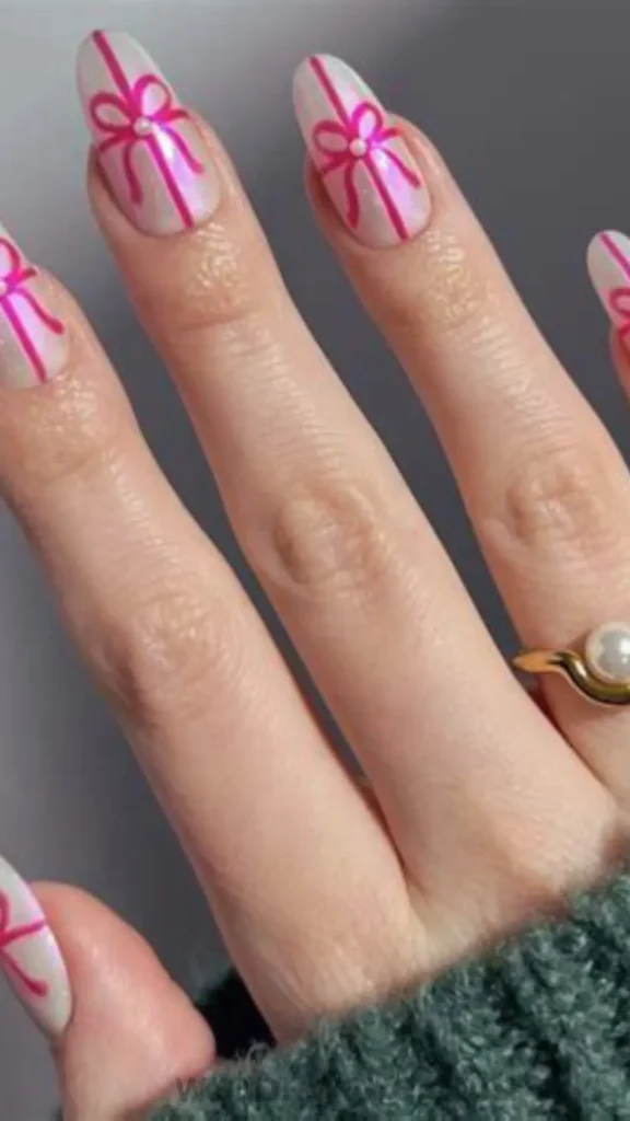 Close-up of almond-shaped gel nails featuring a pearlescent white chrome base decorated with bright magenta pink painted ribbon bows and tiny pearl center studs. A vibrant visual example of ribbon bow nail art and coquette manicure trends.
