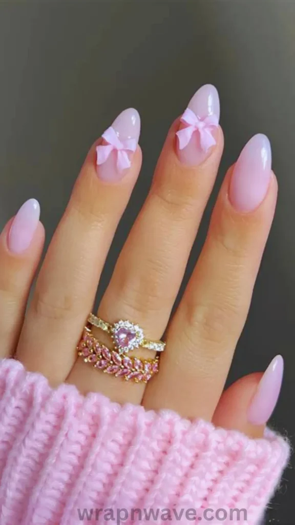 Close-up of almond-shaped gel nails in a sheer, milky pastel pink, decorated with 3D sculpted baby pink ribbon bows on two accent nails and paired with gold and pink gemstone rings. A perfect visual example of coquette nails and 3D bow manicure trends.